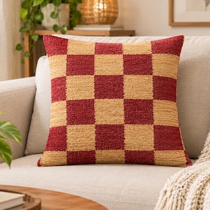 May include: A square throw pillow featuring a textured checkerboard pattern in alternating deep red and tan squares. The fabric has a visible woven, looped pile construction, creating a tactile surface across the entire front of the cushion.