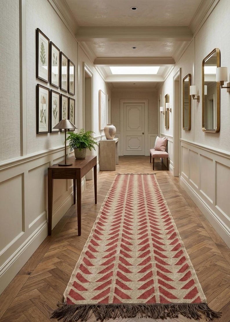 A red and cream herringbone pattern runner rug with fringed ends on a wood floor in a hallway with white wainscoting.