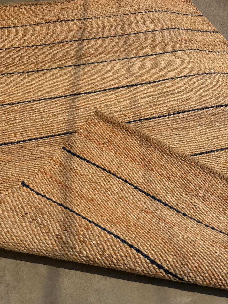 Pode incluir: Um tapete retangular tecido com fibras naturais de cor castanho-claro, apresentando uma s&eacute;rie de riscas finas e paralelas em azul-marinho escuro que percorrem a superf&iacute;cie horizontalmente. O tapete est&aacute; dobrado num dos cantos.