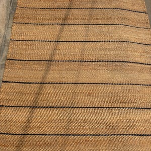 Pode incluir: Um tapete retangular feito de fibras naturais tecidas em tom castanho, apresentando uma s&eacute;rie de riscas horizontais finas em azul-marinho escuro espa&ccedil;adas uniformemente por toda a superf&iacute;cie, criando um padr&atilde;o linear simples no material.