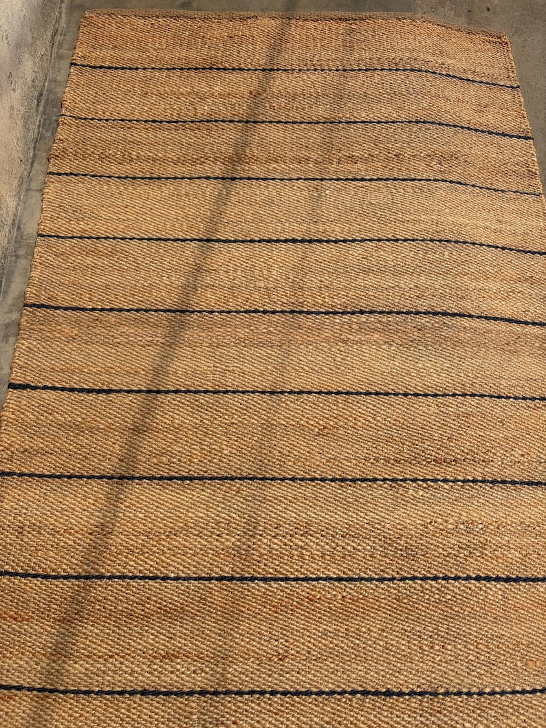 Pode incluir: Um tapete retangular feito de fibras naturais tecidas em tom castanho, apresentando uma s&eacute;rie de riscas horizontais finas em azul-marinho escuro espa&ccedil;adas uniformemente por toda a superf&iacute;cie, criando um padr&atilde;o linear simples no material.