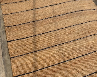 Può includere: Un tappeto rettangolare intrecciato in fibre di juta color marrone naturale, caratterizzato da una serie di sottili strisce orizzontali nere parallele, distanziate uniformemente su tutta la superficie. La trama è grossolana e intrecciata.
