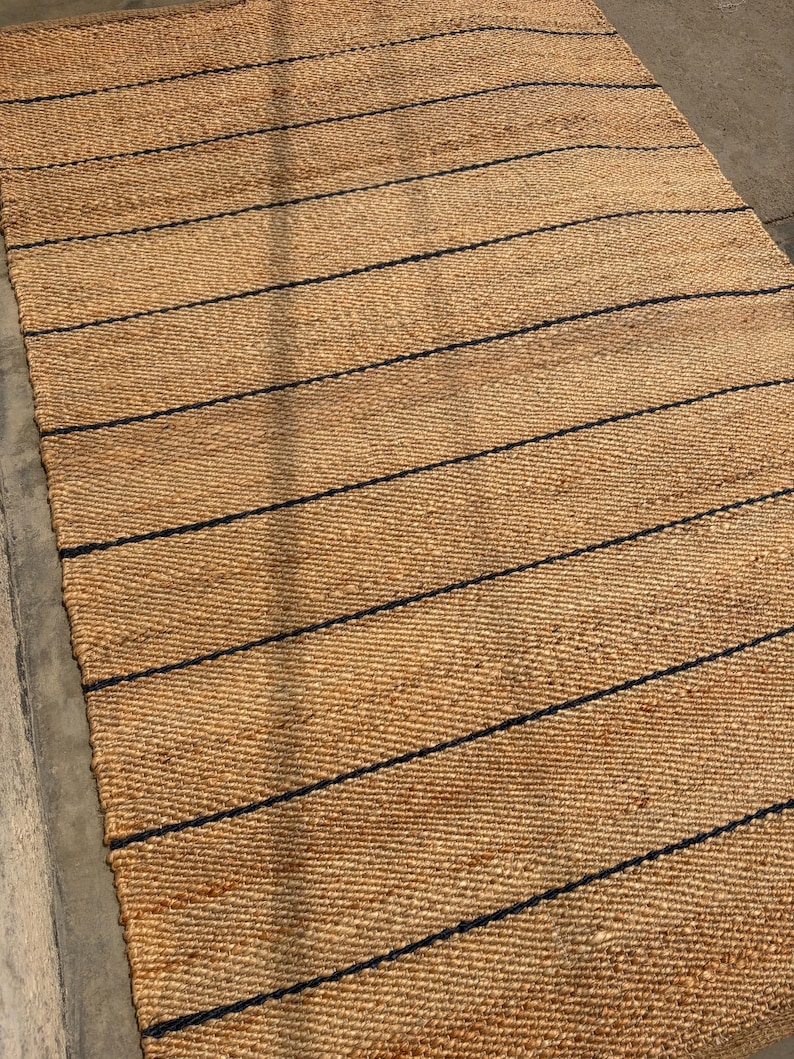 Pode incluir: Um tapete retangular tecido com fibras naturais de cor castanha, apresentando uma s&eacute;rie de riscas horizontais finas, paralelas e de cor azul-marinho escuro, espa&ccedil;adas uniformemente por toda a superf&iacute;cie, criando um padr&atilde;o linear e texturado.
