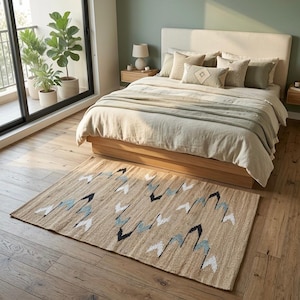 Op de afbeelding: Een slaapkamer met een beige tapijt met een geometrisch patroon van zwarte, witte en lichtblauwe vormen. Het tapijt ligt op een houten vloer voor een bed met een beige hoofdbord en beddengoed. Een balkon met potplanten is zichtbaar.