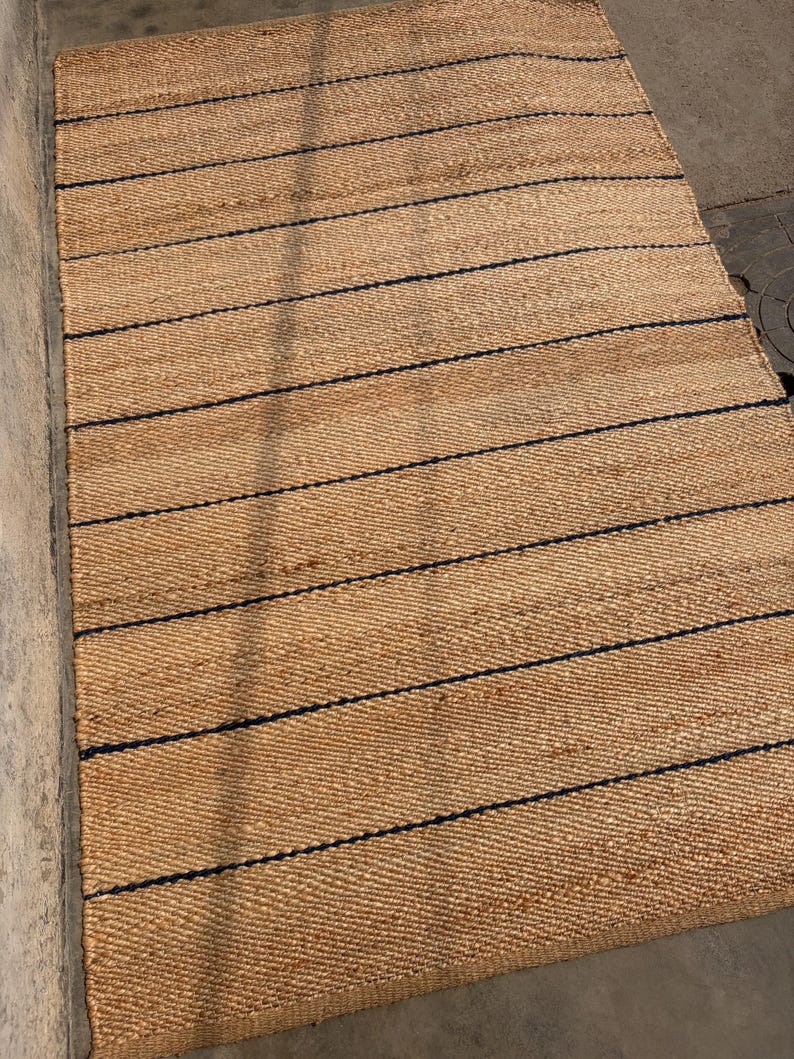 Pode incluir: Um tapete retangular tecido com fibras naturais de cor castanha, apresentando uma s&eacute;rie de riscas horizontais paralelas finas de cor azul-escura, espa&ccedil;adas uniformemente pela superf&iacute;cie. A textura &eacute; grossa e entran&ccedil;ada, criando um padr&atilde;o consistente.