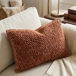 May include: A rectangular throw cushion in a deep terracotta orange colour, featuring a dense, textured surface covered in small, raised pom-pom-like spheres. The cushion has a visible stitched border edge and is resting on a light beige sofa.