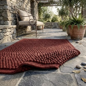 May include: A rectangular rug featuring a deep red color and a textured surface composed of numerous small, rounded, pebble-like spheres. The rug is partially folded at one corner, revealing its thick, dense construction and uniform dark red hue.