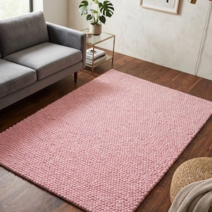 Può includere: Un tappeto rettangolare rosa chiaro con una superficie strutturata e nodosa. Il tappeto è posizionato su un pavimento in legno, con un divano grigio parzialmente visibile sullo sfondo. Il tappeto aggiunge un elemento morbido e caldo all'arredamento della stanza.