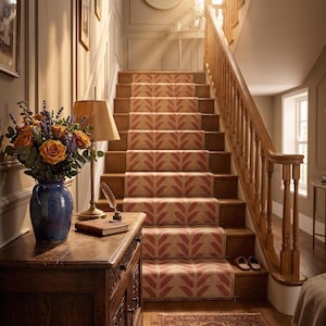 A red and cream herringbone pattern runner rug on wooden stairs with a light wood banister, with a lamp and flowers on a chest.