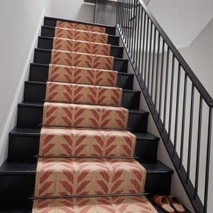A red and cream herringbone pattern runner rug on black painted stairs with a black metal banister, with two brown slippers at the bottom.