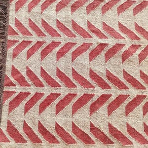 A close-up, overhead view of a red and cream herringbone pattern rug with visible fringe on the left edge.