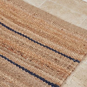 Pode incluir: Um tapete retangular feito de fibras naturais tecidas em tom castanho, com uma textura distinta e duas riscas paralelas azul-marinho escuro que correm diagonalmente pela superf&iacute;cie. O tapete apresenta uma constru&ccedil;&atilde;o entran&ccedil;ada e grossa.