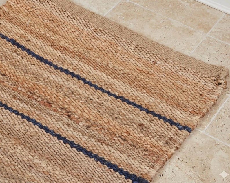 Pode incluir: Um tapete retangular feito de fibras naturais tecidas em tom castanho, com uma textura distinta e duas riscas paralelas azul-marinho escuro que correm diagonalmente pela superf&iacute;cie. O tapete apresenta uma constru&ccedil;&atilde;o entran&ccedil;ada e grossa.
