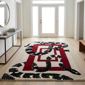 Può includere: Un tappeto rettangolare con sfondo color crema e un audace design a cornice geometrica rosso scuro. Un grande motivo a serpente con un motivo bianco e nero è intrecciato sulla superficie, sovrapponendosi ai bordi rettangolari rossi.