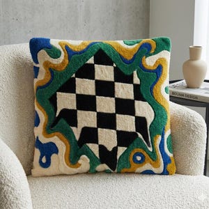 Puede incluir: Un cojín cuadrado decorativo con un centro a cuadros blanco y negro. El cojín tiene un fondo color crema con un borde ondulado en azul, verde y amarillo. El cojín está sobre una silla color crema.