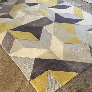 Op de afbeelding: Een rechthoekig vloerkleed met een geometrisch patroon van driehoeken en polygonen in de kleuren mosterdgeel, antracietgrijs, lichtgrijs en crème. Het oppervlak toont een mix van gesneden en geluste texturen door het hele ontwerp heen.