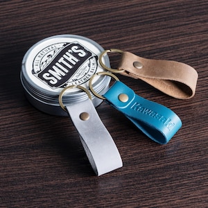 May include: Three leather keychains in gray, tan, and blue with brass rings, next to a tin of Smith's Leather Balm. The blue keychain has the word "Kawasaki" embossed on it. The tin is silver with a black and white label.