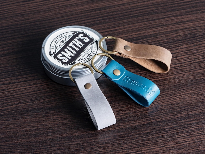 May include: Three leather keychains in gray, tan, and blue with brass rings, next to a tin of Smith's Leather Balm. The blue keychain has the word "Kawasaki" embossed on it. The tin is silver with a black and white label.