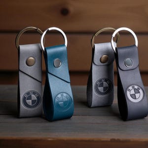 May include: Leather keychains in gray, teal, and black, each featuring the BMW logo. The keychains have a metal ring and a snap closure. The keychains are displayed on a wooden surface.
