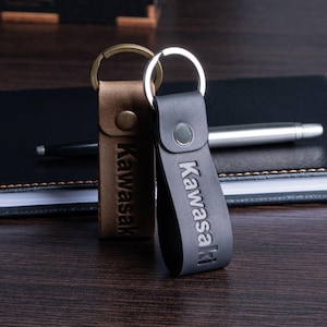 May include: Two leather keychains with the word "Kawasaki" embossed on them. One keychain is brown with a gold-colored ring, and the other is black with a silver-colored ring. They are resting on a dark wooden surface next to a black notebook and pen.