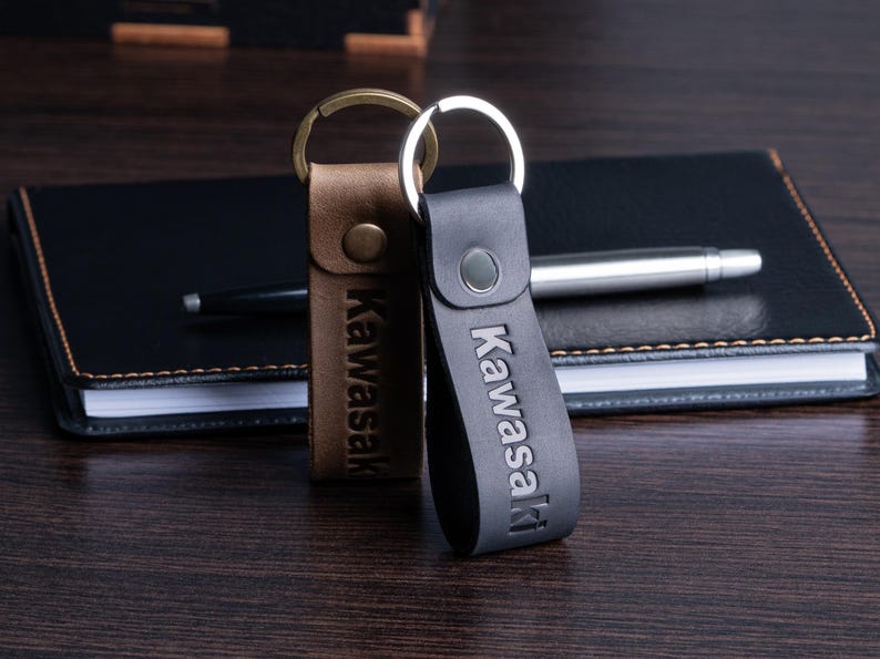 May include: Two leather keychains with the word "Kawasaki" embossed on them. One keychain is brown with a gold-colored ring, and the other is black with a silver-colored ring. They are resting on a dark wooden surface next to a black notebook and pen.