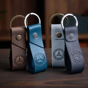 Könnte beinhalten: Vier Leder-Schlüsselanhänger in Braun, Türkis, Grau und Marineblau. Jeder Schlüsselanhänger hat einen Metallring und einen Druckknopfverschluss. Das Mercedes-Benz-Logo ist auf jedem Lederriemen geprägt.