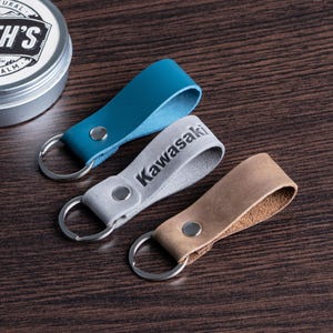 May include: Three leather keychains in teal, gray with "Kawasaki" printed, and tan. Each keychain has a silver ring and a metal rivet. A tin of leather balm is in the background.