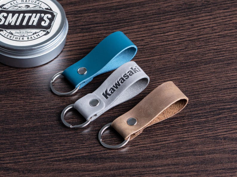 May include: Three leather keychains in teal, gray with "Kawasaki" printed, and tan. Each keychain has a silver ring and a metal rivet. A tin of leather balm is in the background.
