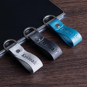 May include: Three leather keychains in gray, black, and teal, each with a silver keyring and the embossed word "Kawasaki." The keychains are arranged on a dark wooden surface next to a black leather notebook.