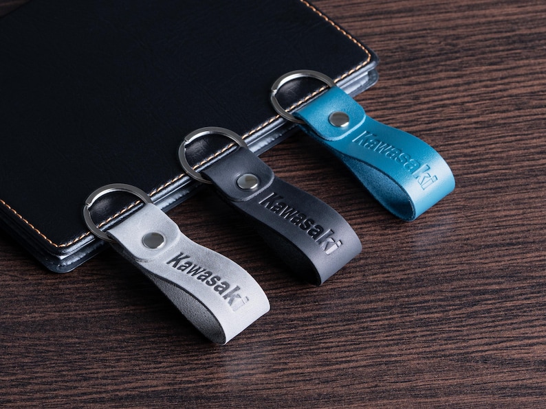 May include: Three leather keychains in gray, black, and teal, each with a silver keyring and the embossed word "Kawasaki." The keychains are arranged on a dark wooden surface next to a black leather notebook.