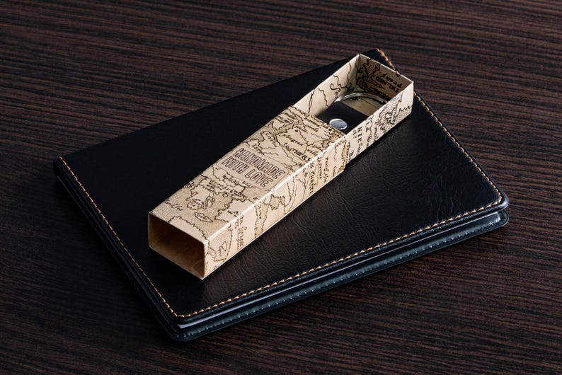 May include: A black leather notebook with a stitched border, topped with a rectangular box. The box is beige with a world map design and the words "HANDMADE WITH LOVE". Inside the box is a silver-toned object.