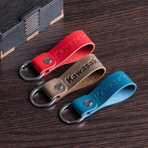 May include: Three leather keychains in red, tan, and blue, each with the embossed word "Kawasaki". The keychains have silver-colored key rings and metal snap closures. They are arranged on a dark wood surface.