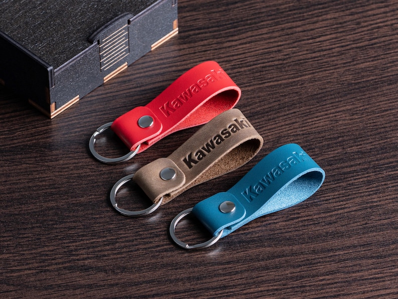 May include: Three leather keychains in red, tan, and blue, each with the embossed word "Kawasaki". The keychains have silver-colored key rings and metal snap closures. They are arranged on a dark wood surface.