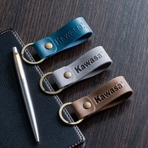 May include: Three leather keychains in teal, gray, and brown, each embossed with the name "Kawasaki". The keychains have brass-colored key rings and rivets. A silver pen and a dark blue leather notebook are also in the image.