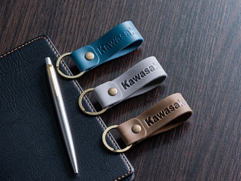 May include: Three leather keychains in teal, gray, and brown, each embossed with the name "Kawasaki". The keychains have brass-colored key rings and rivets. A silver pen and a dark blue leather notebook are also in the image.