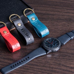 May include: A collection of leather keychains in red, gray, and teal, each with "Kawasaki" embossed. A dark gray smartwatch with a black bezel and a dark blue band is also present. The items are arranged on a wooden surface.