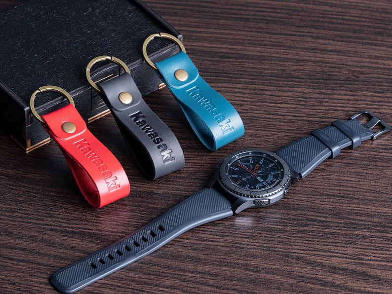May include: A collection of leather keychains in red, gray, and teal, each with "Kawasaki" embossed. A dark gray smartwatch with a black bezel and a dark blue band is also present. The items are arranged on a wooden surface.