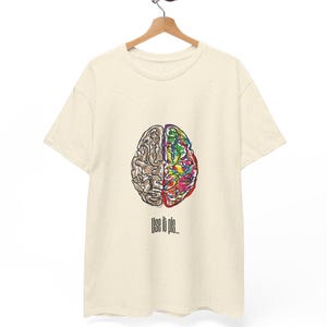 Op de afbeelding: Crèmekleurig T-shirt met een gedetailleerde hersenafbeelding. De hersenen zijn verdeeld, één kant bruin, de andere kleurrijk. De tekst "Use it pls..." staat eronder. Het T-shirt hangt aan een houten hanger.