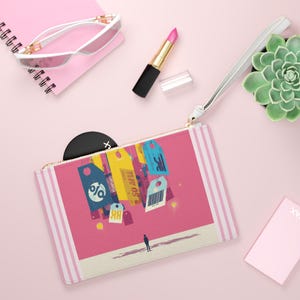 Könnte beinhalten: Eine rosa Clutch mit weißer Handschlaufe, mit einer Grafik von schwebenden Tags und einer Person. Die Clutch liegt neben einem rosa Notizblock, einer Sonnenbrille, Lippenstift und einer Sukkulentenpflanze. Die Tags haben Text und verschiedene Formen.
