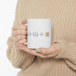 May include: White ceramic coffee mug featuring black graphics of a computer key, the word "Alt", a plus sign, and the word "Coffee" in a wooden-style font. The mug is held by a person.