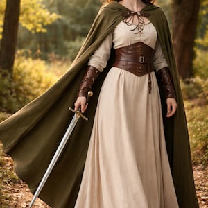 May include: A woman in a forest setting wearing a medieval-style outfit. The ensemble includes a long beige dress, a brown leather corset, and a green cape. She holds a silver sword, embodying a fantasy or historical theme.