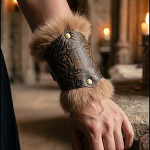 Könnte beinhalten: Ein brauner Lederarmreif mit geprägten Blumen- und Tiermotiven, besetzt mit hellbraunem Fell. Der Armreif wird mit zwei goldfarbenen Knöpfen befestigt. Das Bild zeigt den Armreif am Arm einer Person.