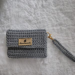 Puede incluir: Un monedero gris hecho a mano con ganchillo, con una correa de muñeca y un cierre dorado. El monedero tiene una pequeña etiqueta rectangular que dice "handmade". El monedero está hecho de un material tejido texturizado.