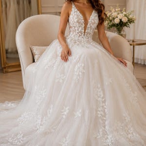 May include: A floor-length ivory wedding dress featuring a deep V-neckline and thin straps. The bodice and full tulle skirt are adorned with intricate white floral lace appliques that cascade down the fabric, creating a textured, layered appearance.