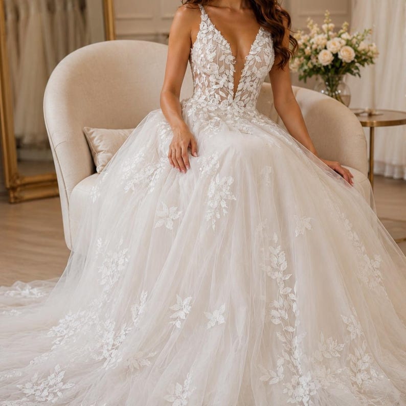 May include: A floor-length ivory wedding dress featuring a deep V-neckline and thin straps. The bodice and full tulle skirt are adorned with intricate white floral lace appliques that cascade down the fabric, creating a textured, layered appearance.