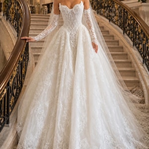 May include: A white strapless ball gown featuring a sweetheart neckline and intricate lace detailing throughout the bodice and full skirt. The dress includes matching long lace sleeves and a long, sheer veil that drapes down the back.