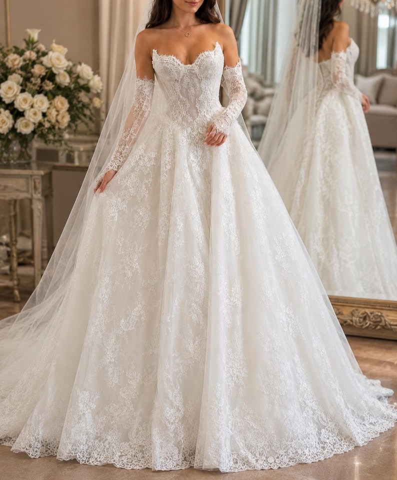 May include: A white strapless ball gown wedding dress featuring an intricate lace bodice and a full, floor-length skirt with lace appliques. The dress includes matching sheer lace long sleeves and a long, flowing veil that drapes over the shoulders.