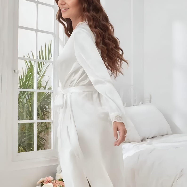 Puede incluir: Una bata de sat&eacute;n blanco con mangas largas y cintur&oacute;n a juego. La bata presenta detalles de encaje en los pu&ntilde;os y el escote. La imagen est&aacute; tomada en una habitaci&oacute;n luminosa con una ventana y una cama al fondo.