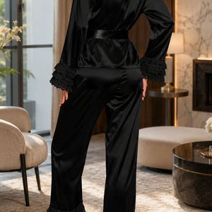 May include: A black two-piece pyjama set consisting of a long-sleeved top and matching trousers. The top features a lace panel across the upper back, a waist tie, and tiered lace ruffles at the cuffs. The trousers have matching tiered lace ruffles at the hems.