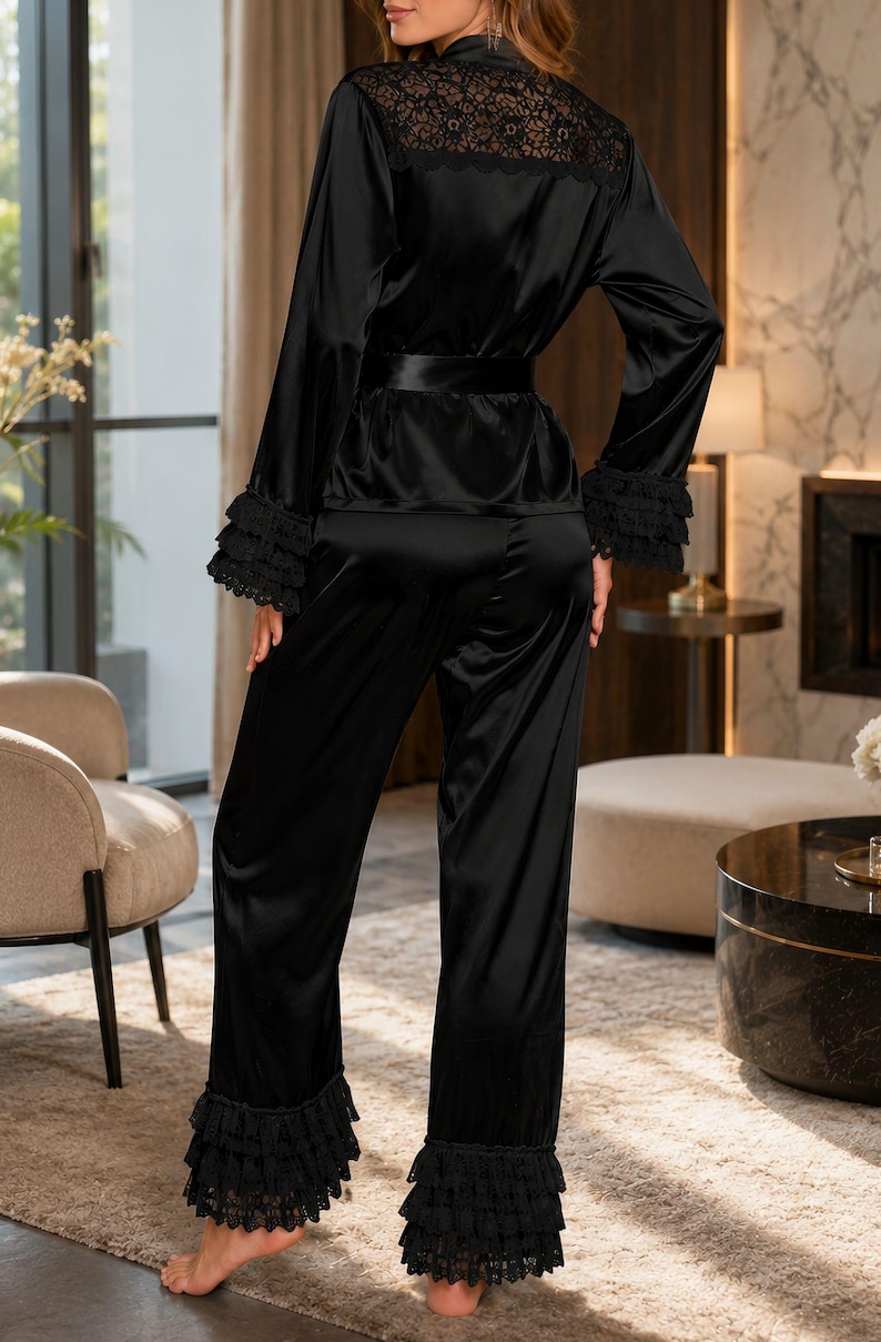 May include: A black two-piece pyjama set consisting of a long-sleeved top and matching trousers. The top features a lace panel across the upper back, a waist tie, and tiered lace ruffles at the cuffs. The trousers have matching tiered lace ruffles at the hems.